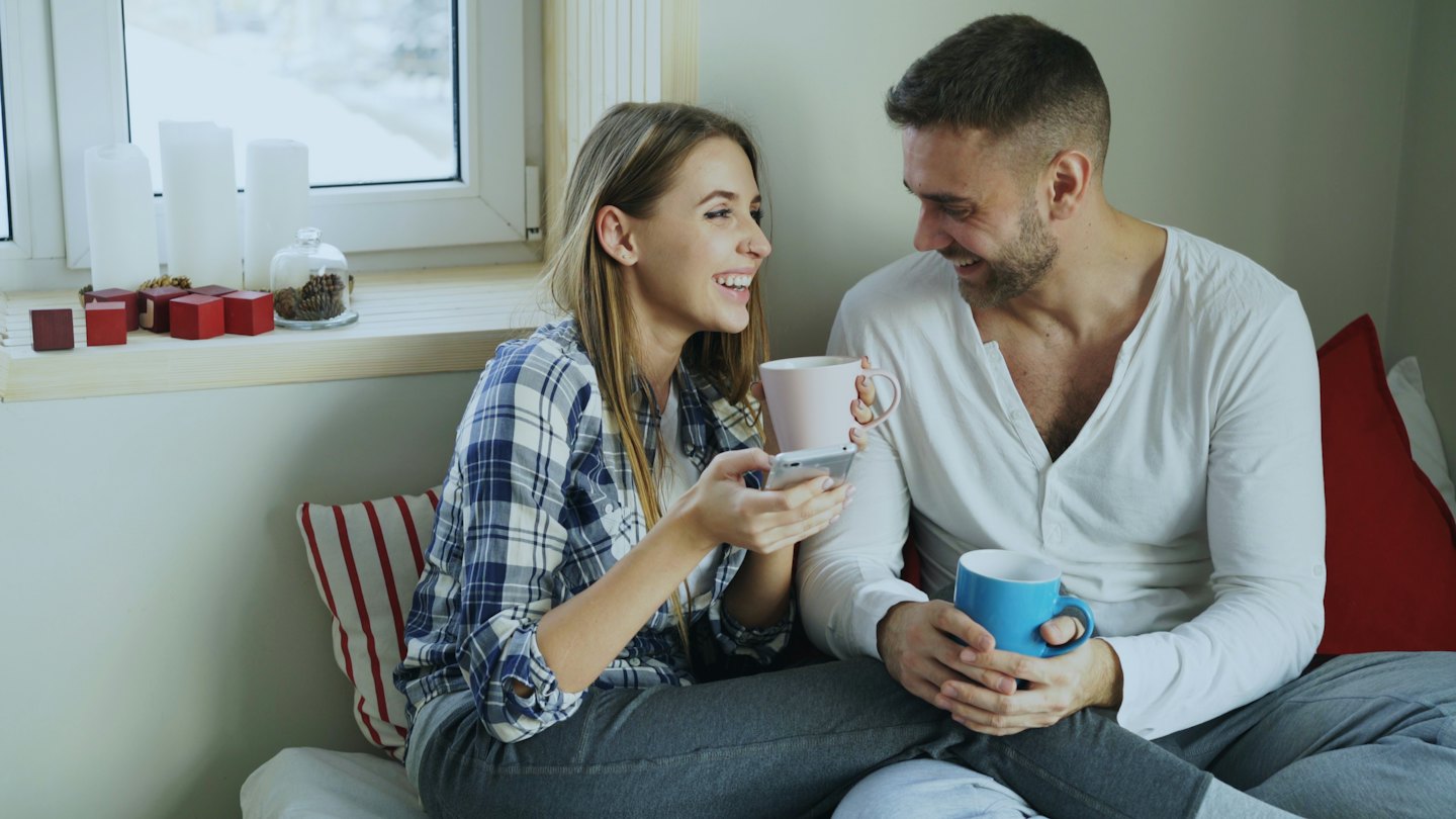 Couple sits by a window laughing while holding coffee mugs.